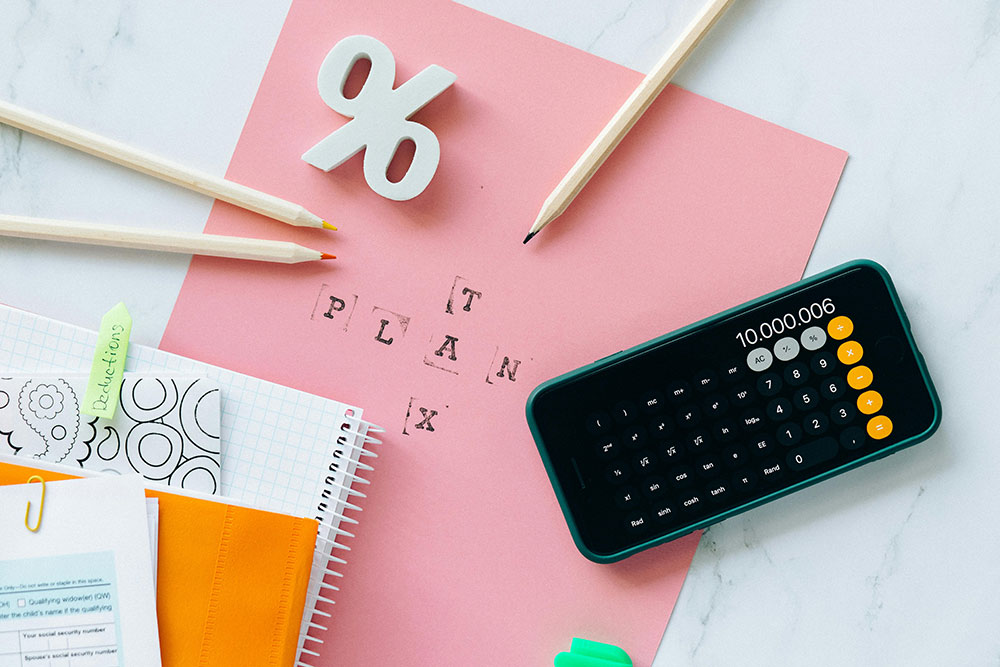 Flat lay of tax planning essentials including a pink sheet with 'TAX PLAN' stamped on it, a smartphone calculator displaying a large number, pencils, and notebooks with a 'Deductions' tab.